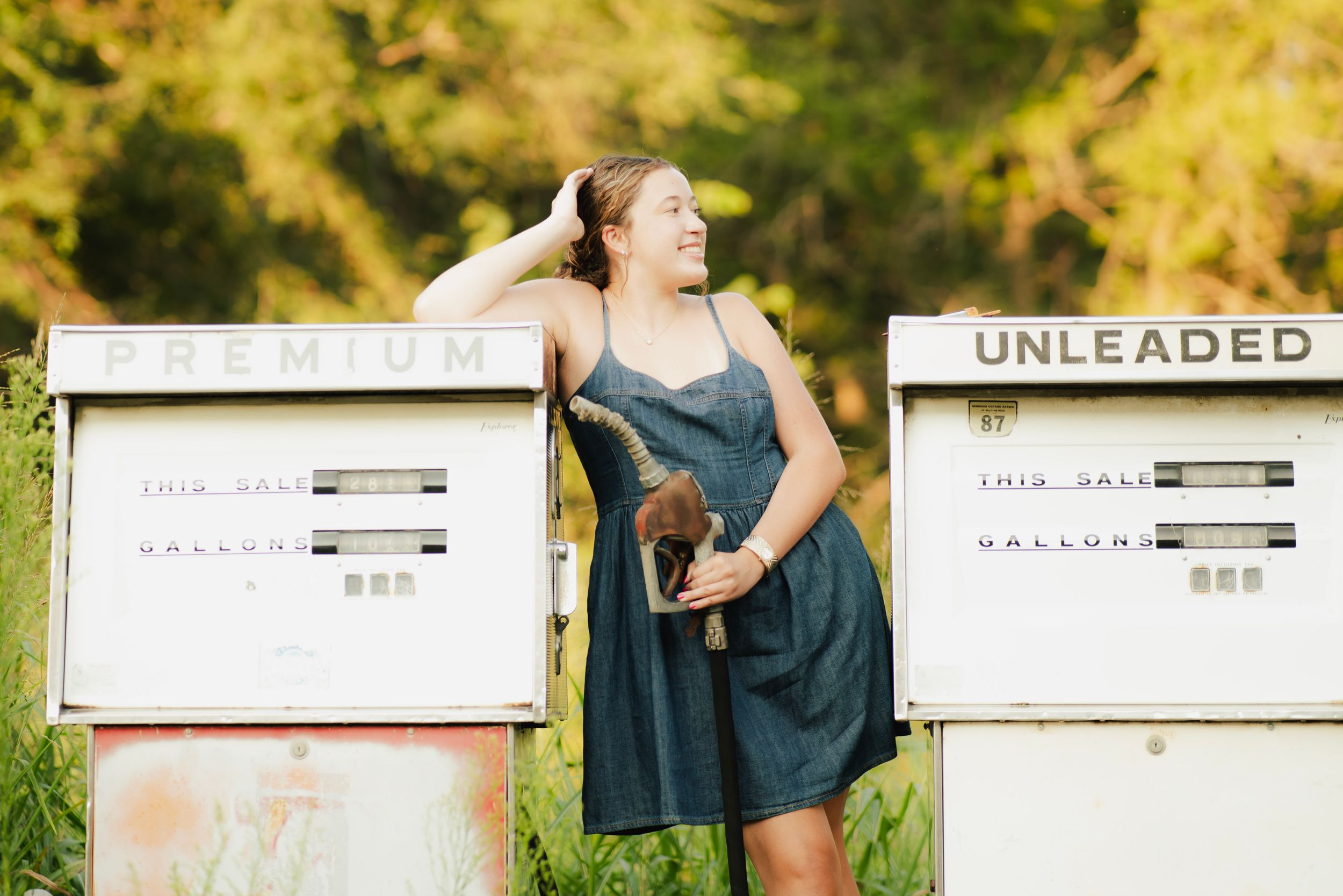 Young woman in denim dress posing between vintage premium and unleaded gas pumps outdoors.