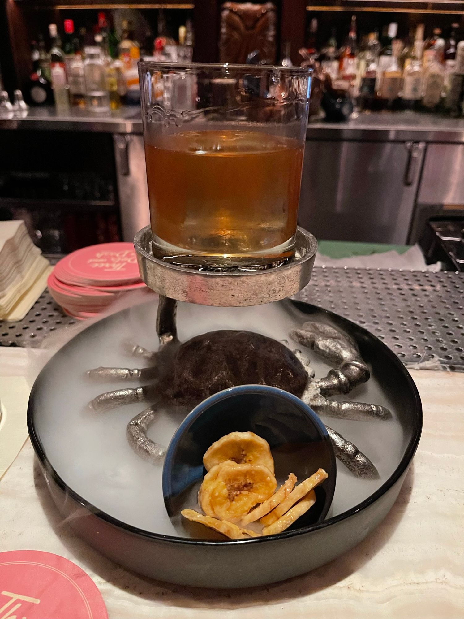 A craft cocktail in a rocks glass on a decorative crab plate with banana chips at a dimly lit bar.