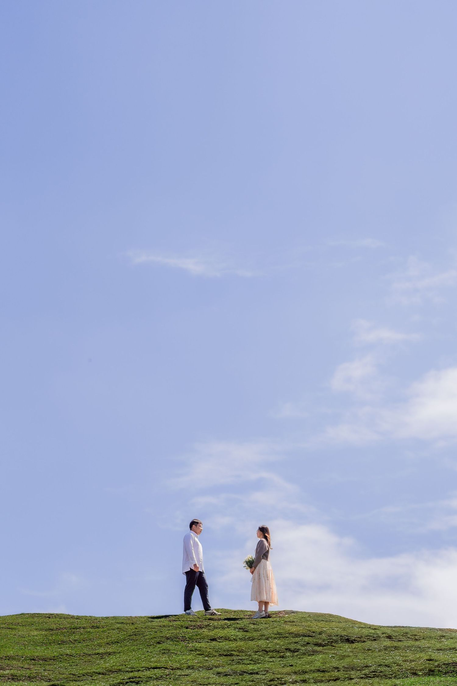 Happy couple sitting on blanket in grass, raising arms in celebration, woman holding white flower bouquet.