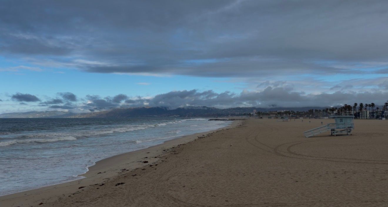 Cloudy beach scene with waves, sandy shore, footprints, and people gathered near the water at dusk.