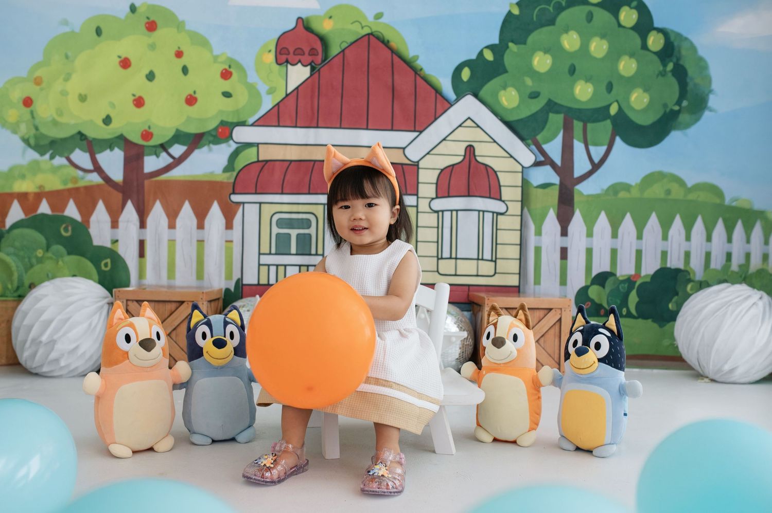 Toddler girl in white dress sits with orange balloon surrounded by Bluey character plush toys against colorful backdrop.