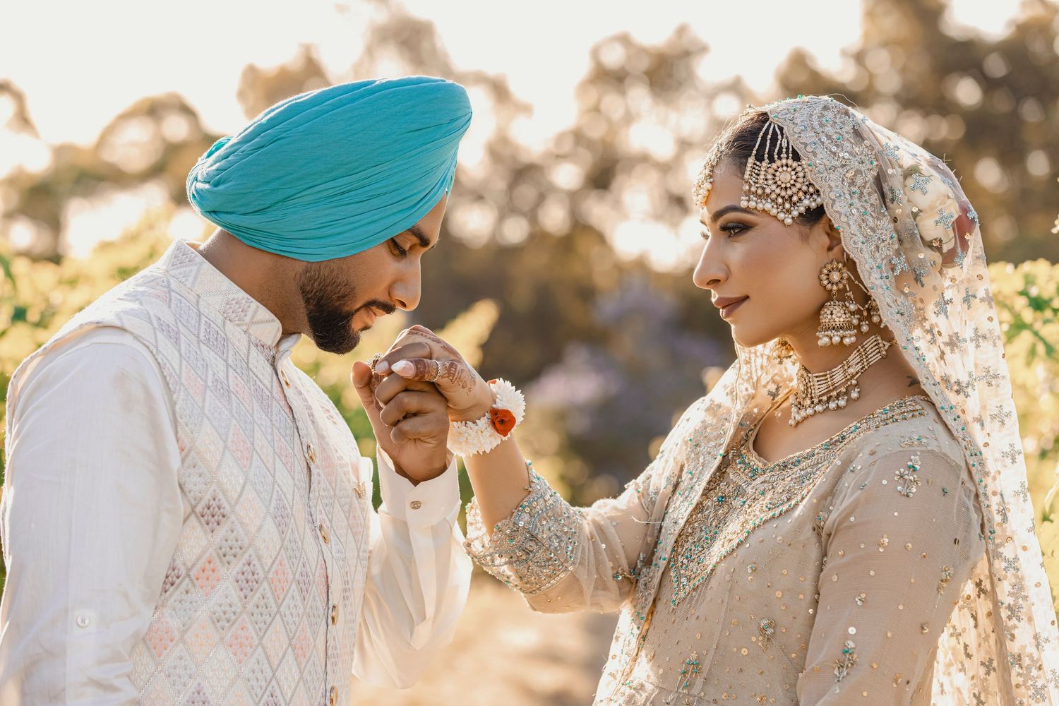 Sikh couple in traditional wedding attire, groom in turban kissing bride's hand outdoors.
