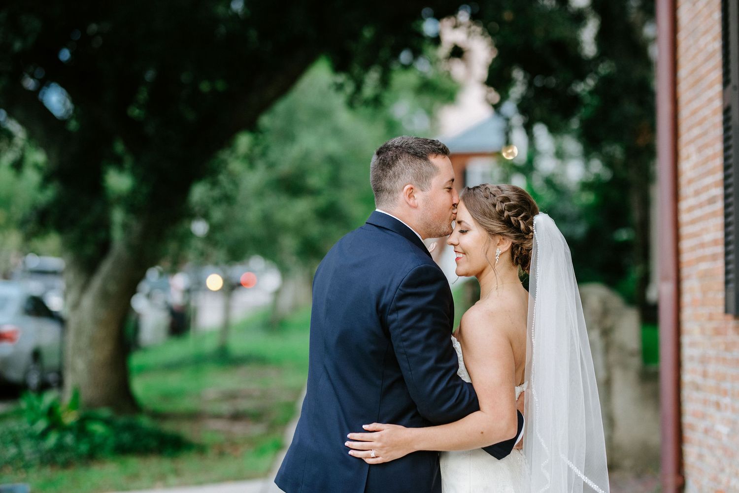 A bride and groom share romantic moments in a garden setting with brick buildings and trees in the background.
