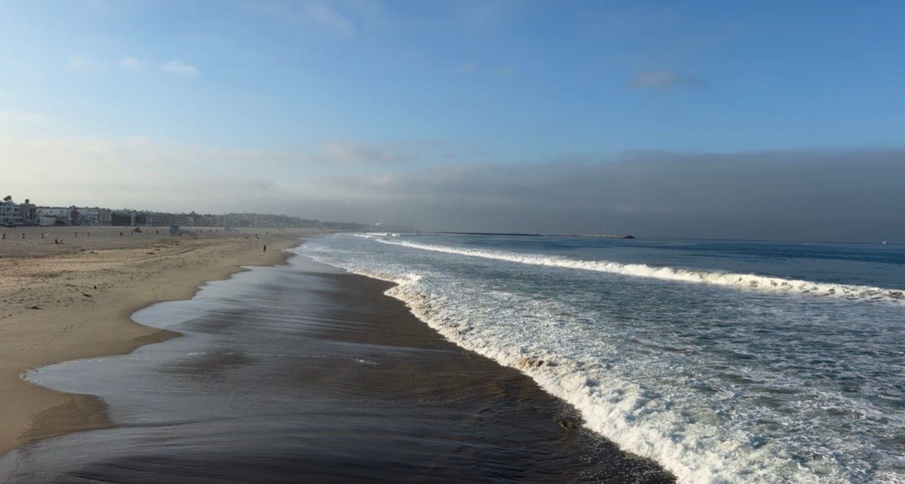 Aerial view of a sandy beach with rolling ocean waves under a blue sky with coastal fog in the distance.