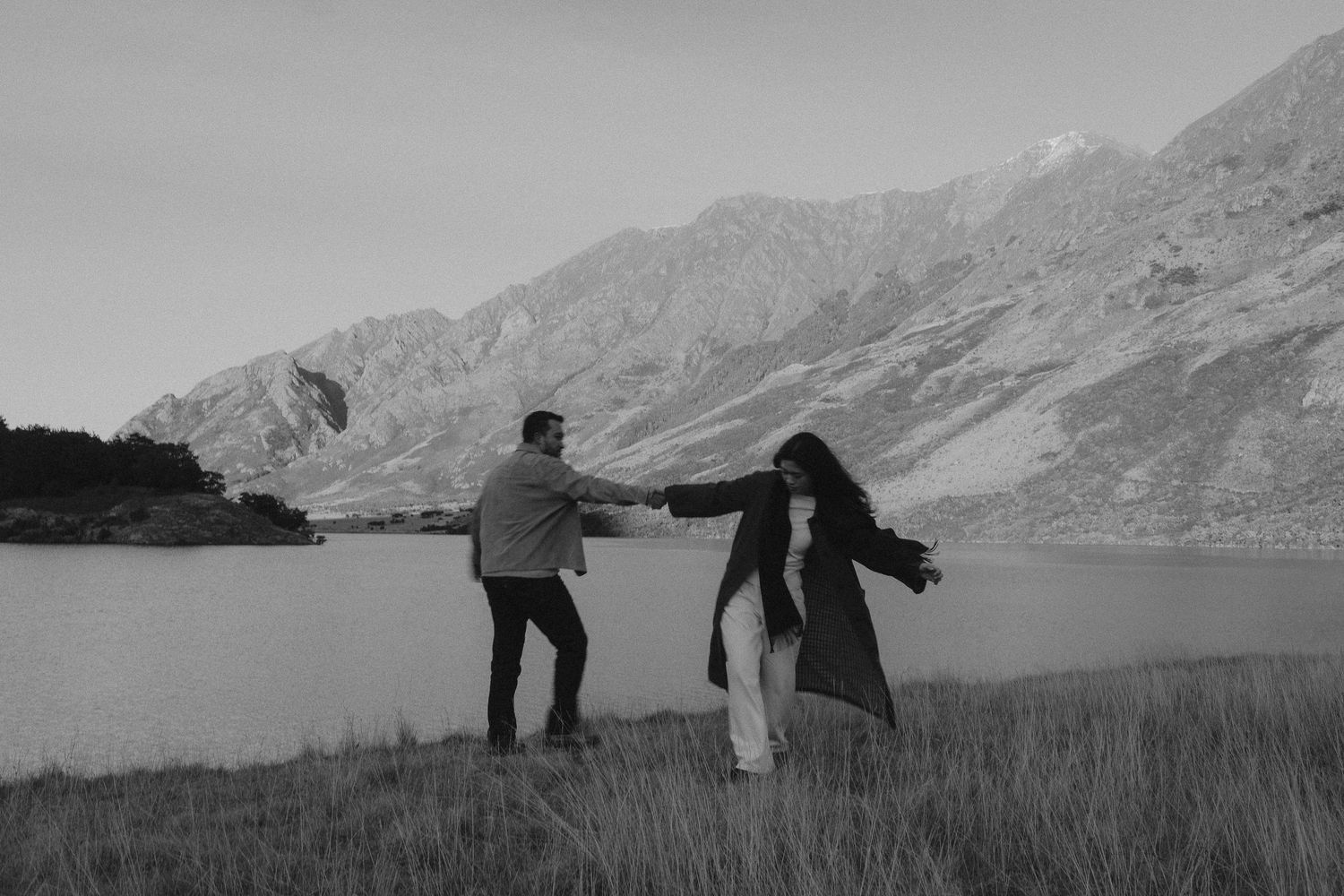 Black and white photo of a couple holding hands by a mountain lake in a grassy field.