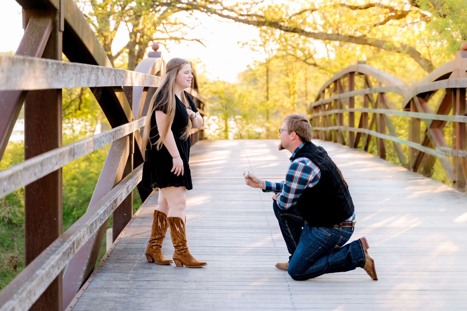 Man proposing on one knee to woman on wooden bridge surrounded by autumn trees at golden hour.