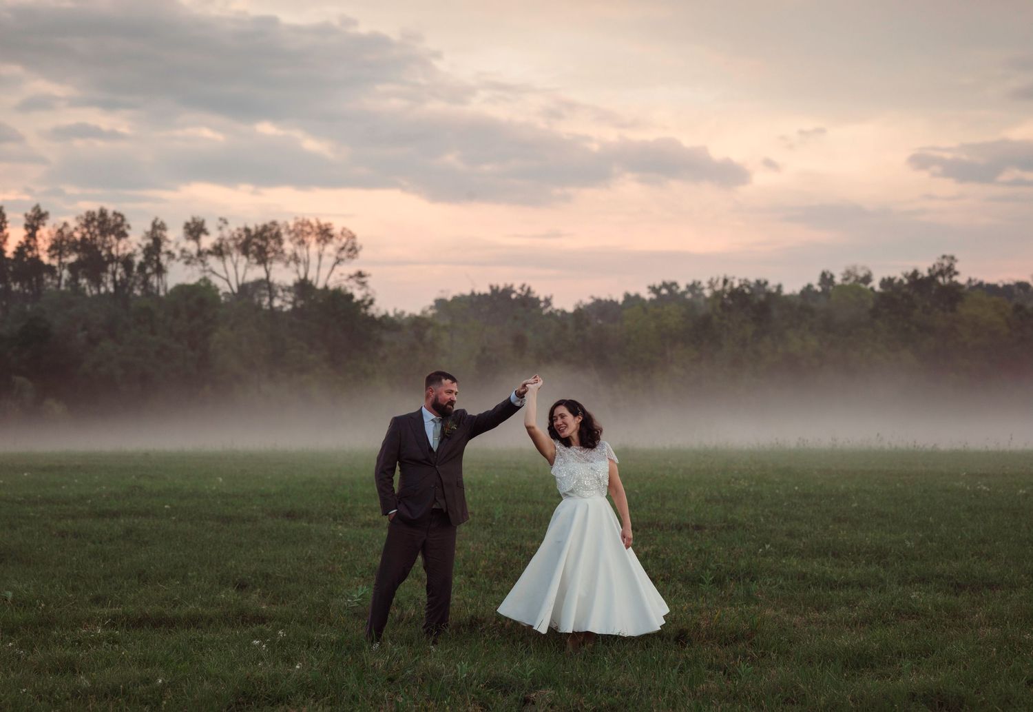Couple shares romantic twirl in lush green meadow field, bride in white gown and groom in black suit on wedding day.