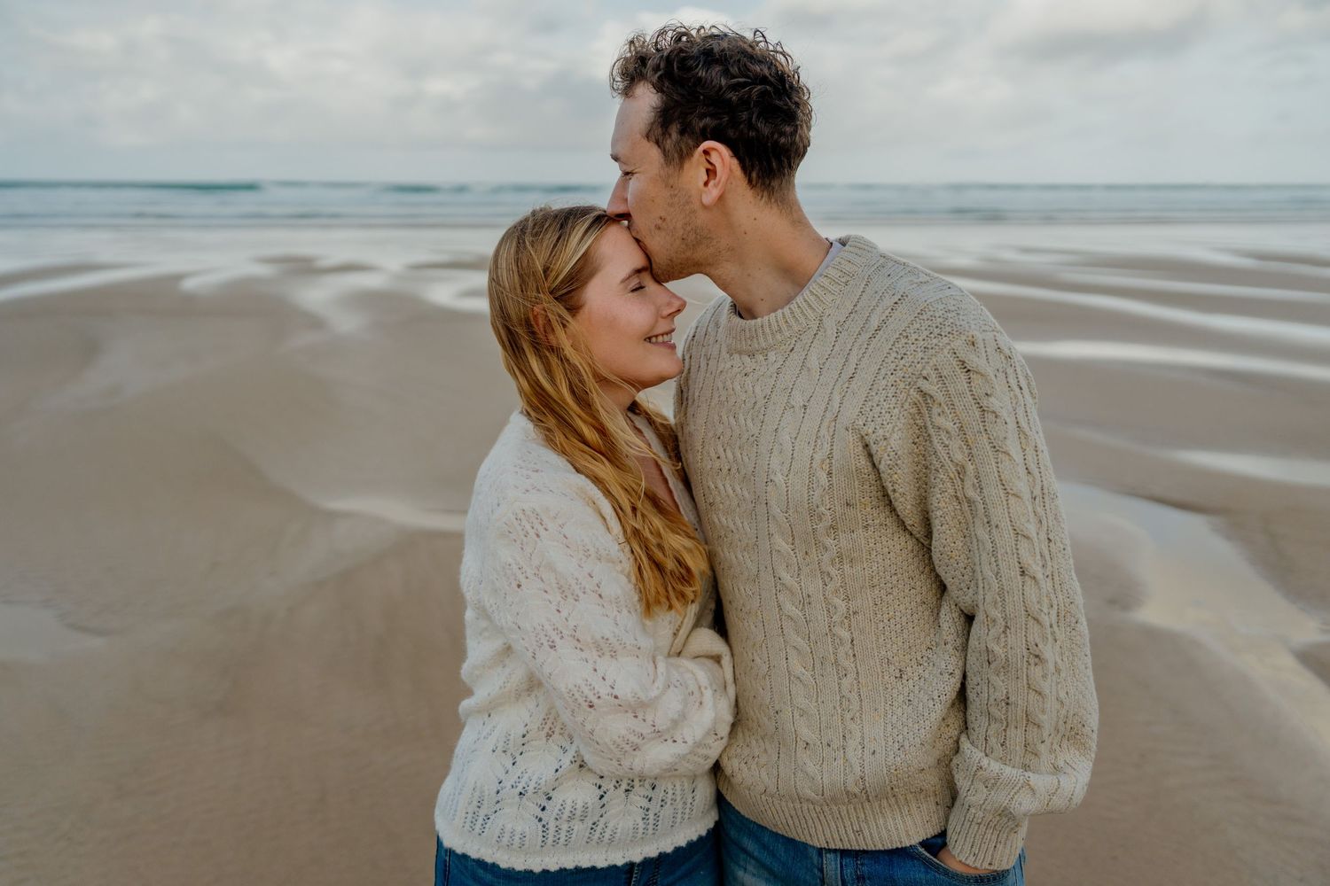 Romantic couple embracing on watergate bay wearing cozy cable-knit sweaters with ocean waves in the background.