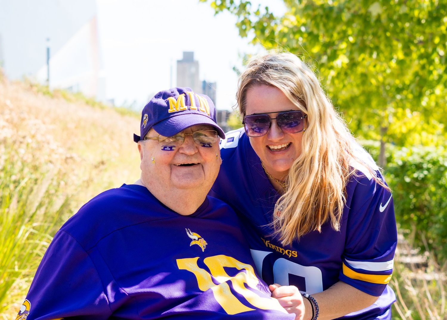 Vikings fan in wheelchair poses by US Bank Stadium Viking ship sculpture on a sunny day.