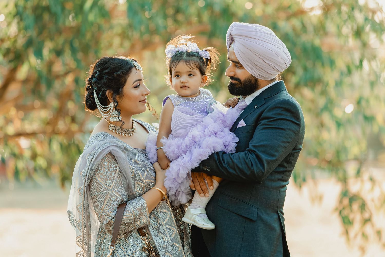 Sikh couple in traditional wedding attire holding their baby girl in a purple dress outdoors.