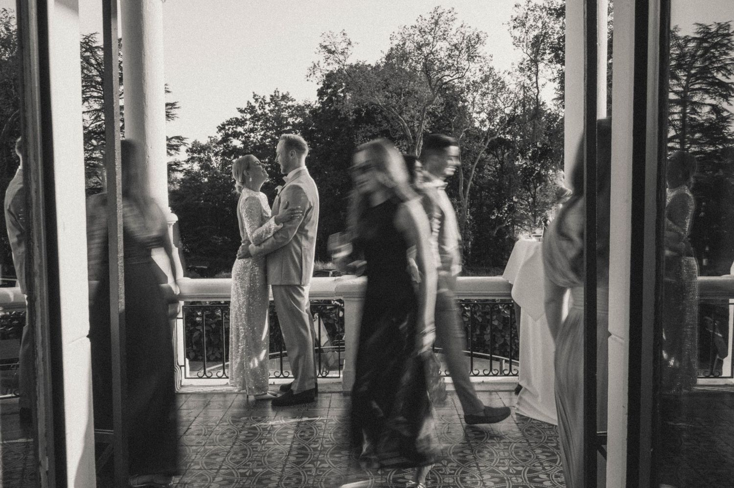 Black and white wedding photo of couple embracing on elegant balcony with blurred guests walking past.
