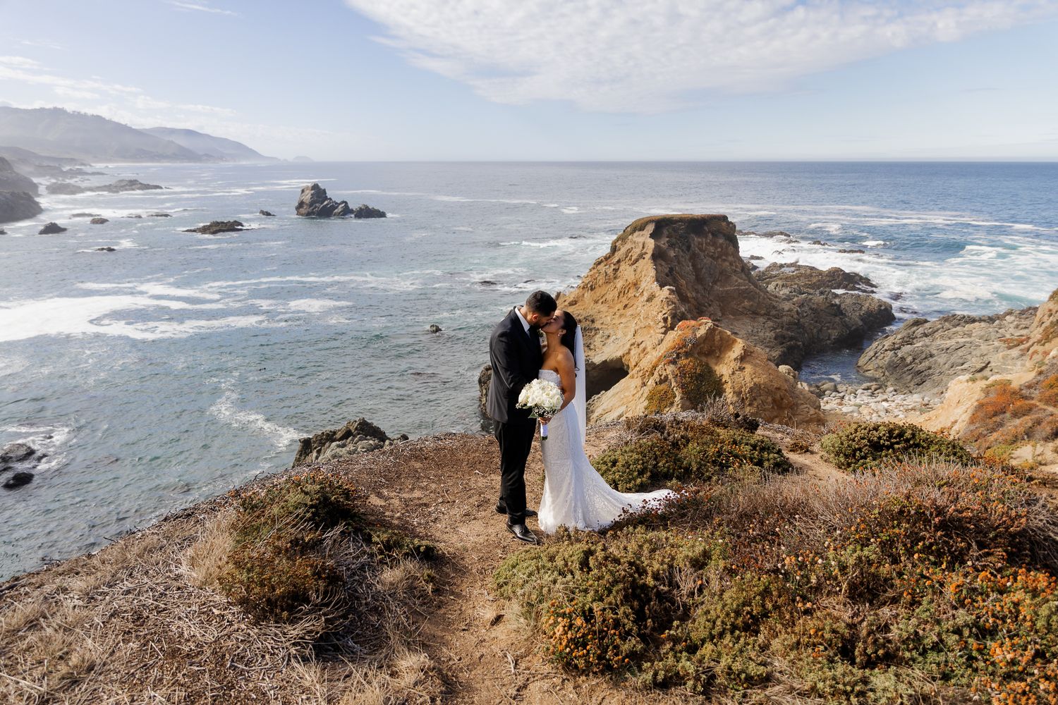 A bride and groom share a romantic kiss on a rugged coastal cliff overlooking the Pacific Ocean on their wedding day.