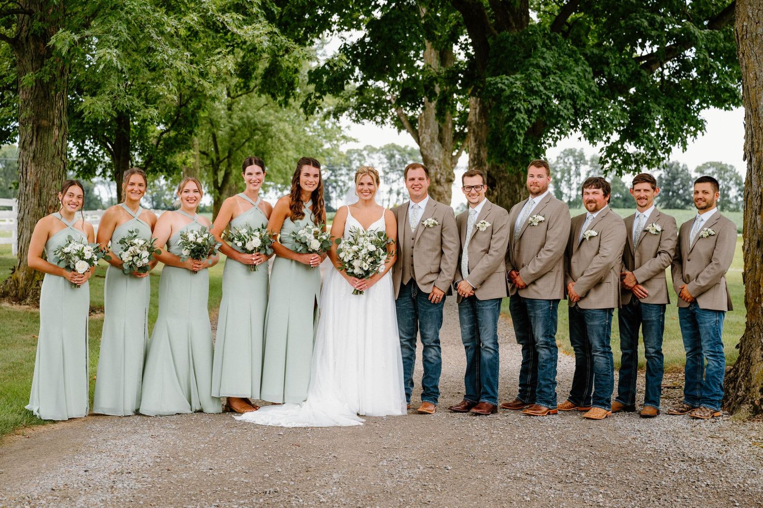 Wedding party poses in sage bridesmaid dresses and tan suits with jeans under large trees.