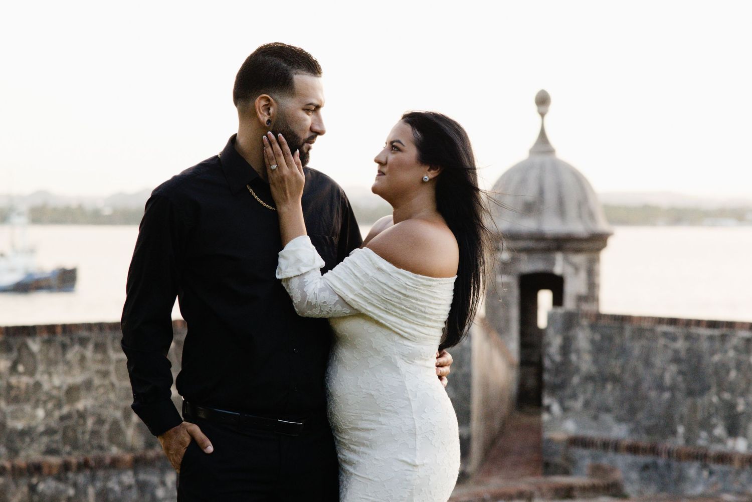 Una pareja se mira junto a un muro de piedra; él viste de negro y ella lleva un vestido blanco.