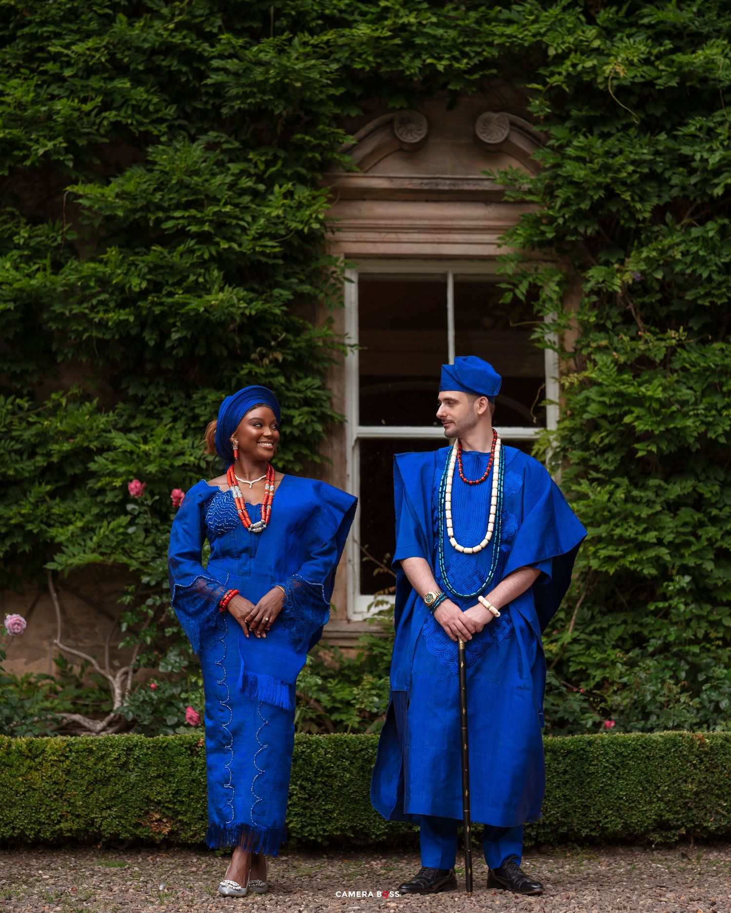 Couple in matching royal blue traditional Nigerian attire standing before ivy-covered stone building.