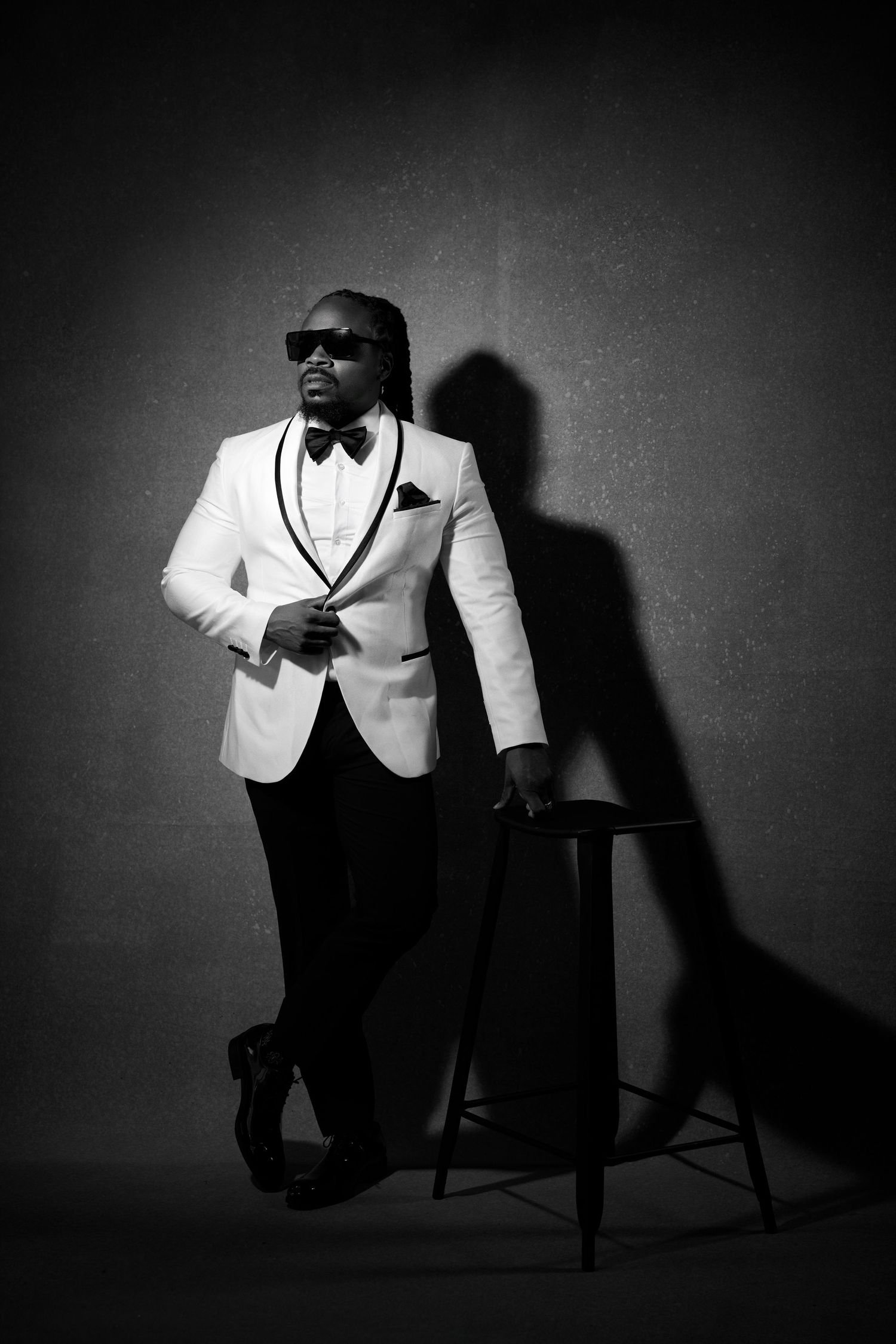 Black and white portrait of a stylish man in white tuxedo jacket, bow tie, and sunglasses leaning on a chair.