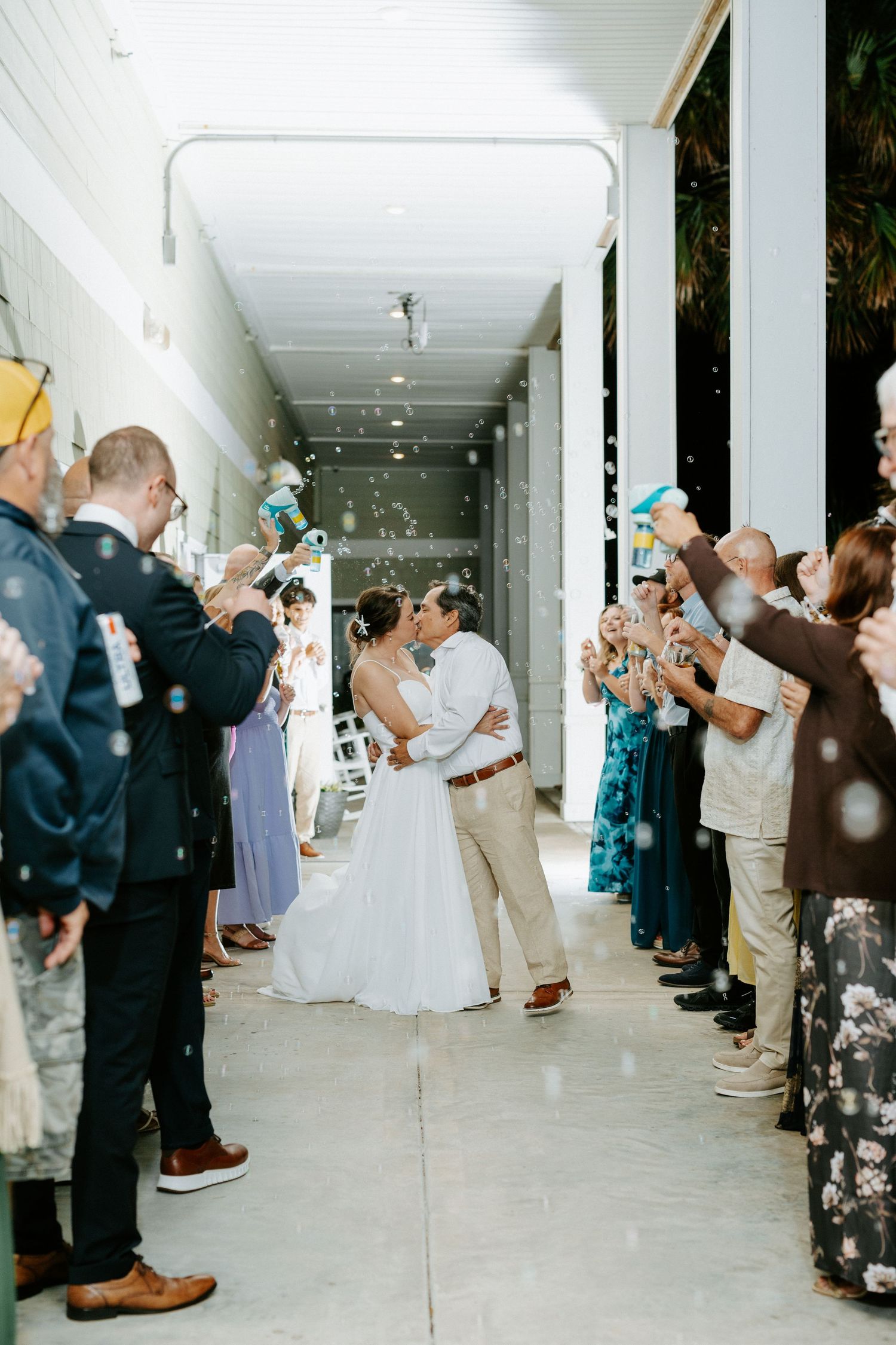 Bride and groom share a kiss while walking through a bubble send-off as wedding guests line a covered outdoor walkway.