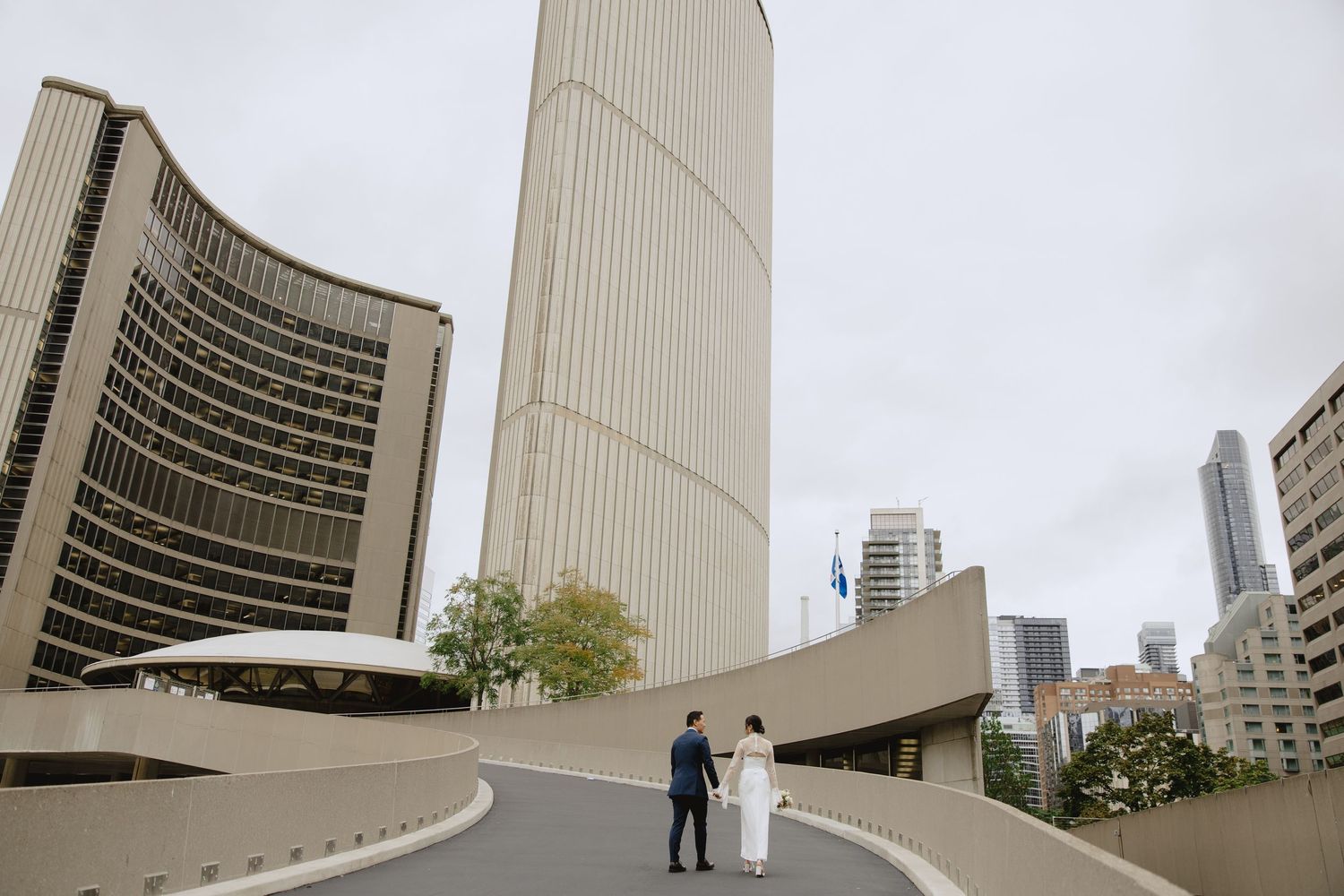 Newlywed couple walking hand in hand along a curved concrete ramp beside modern curved towers under a cloudy sky.