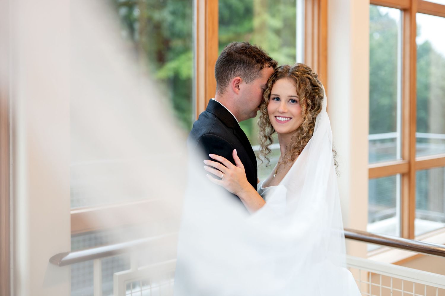 Bride and groom share a romantic kiss in an elegant wedding reception venue with floral centerpieces and large windows.