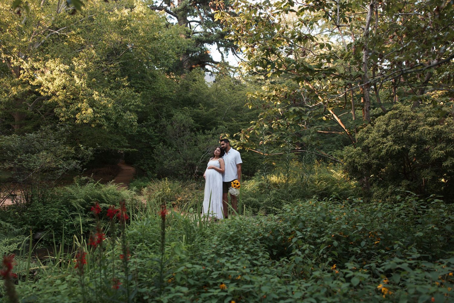 Couple embracing in a lush green garden surrounded by colorful flowers and tall trees at dusk.