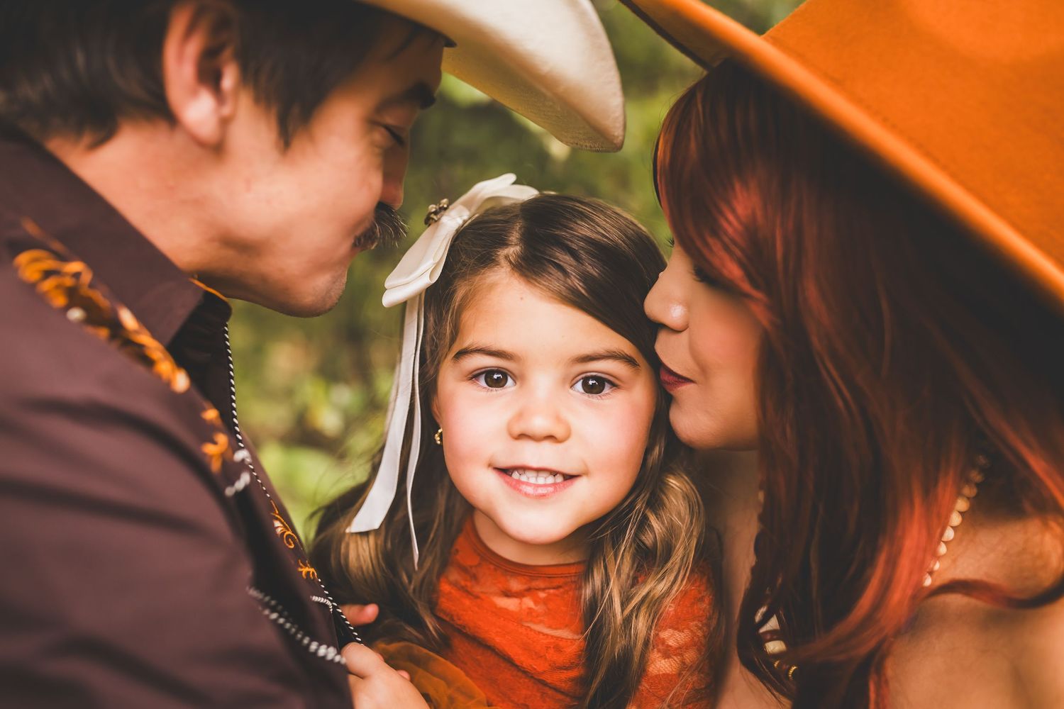 Family in fall costumes walking through woods, woman in red dress and cowboy hat, child in orange tutu dress.
