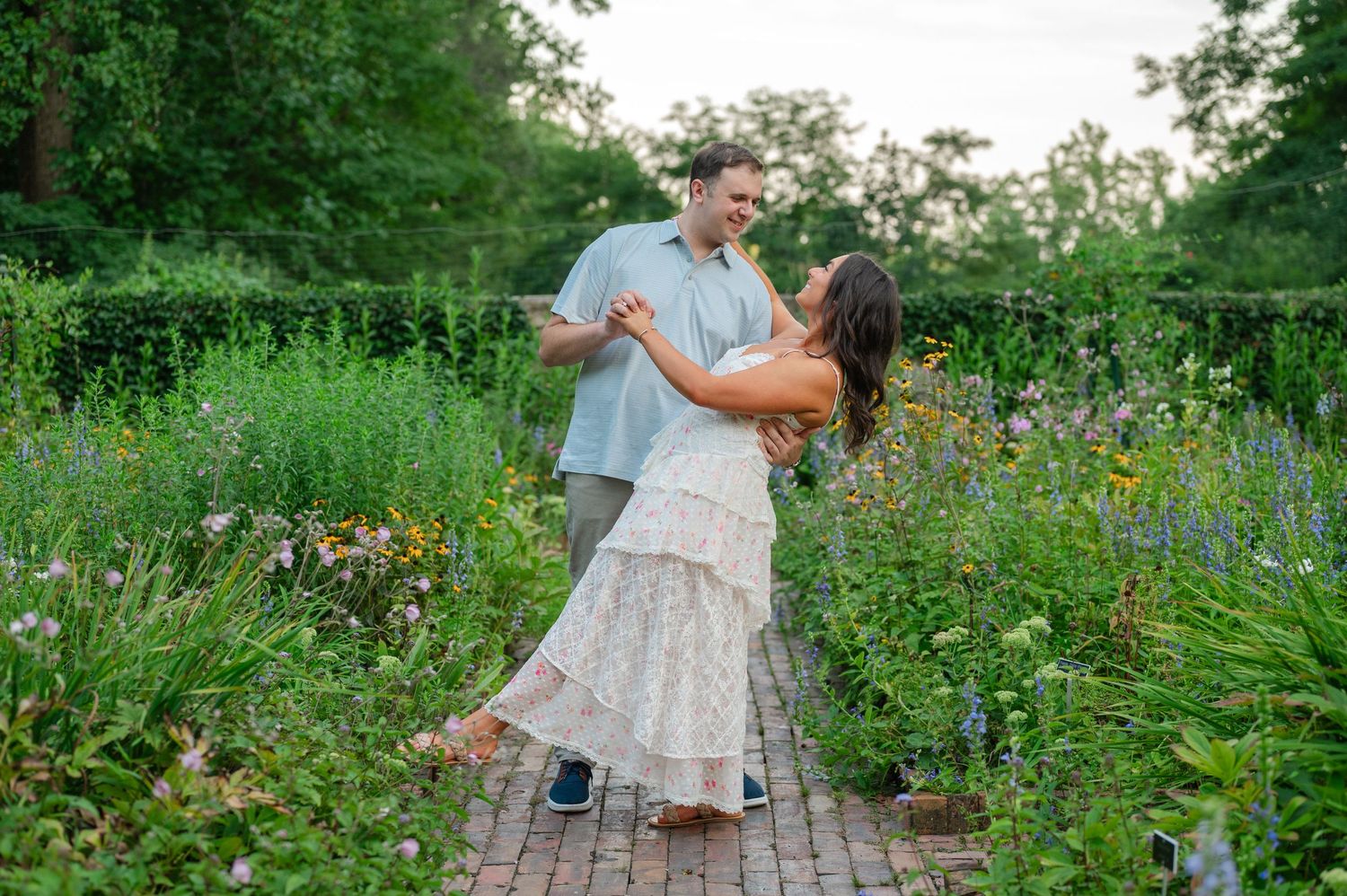 Couple dancing joyfully on a brick path surrounded by lush green gardens and wildflowers in soft natural light.