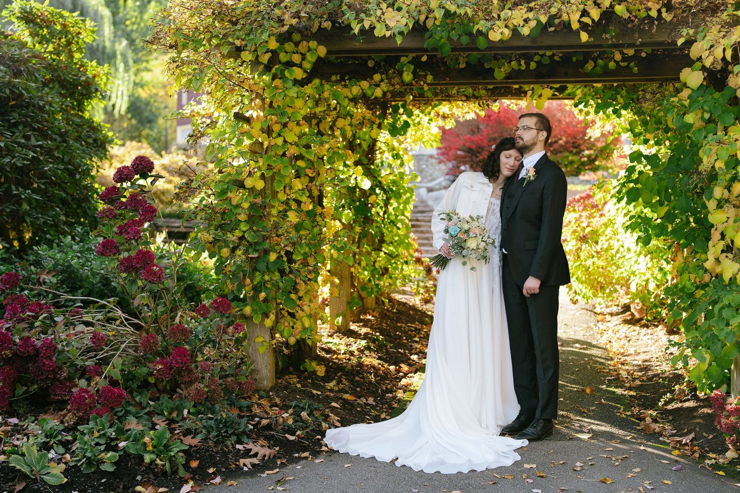 Melissa resting her head on Michael's shoulder under a vine-covered arbor at Deer Lake Park, holding her bouquet.