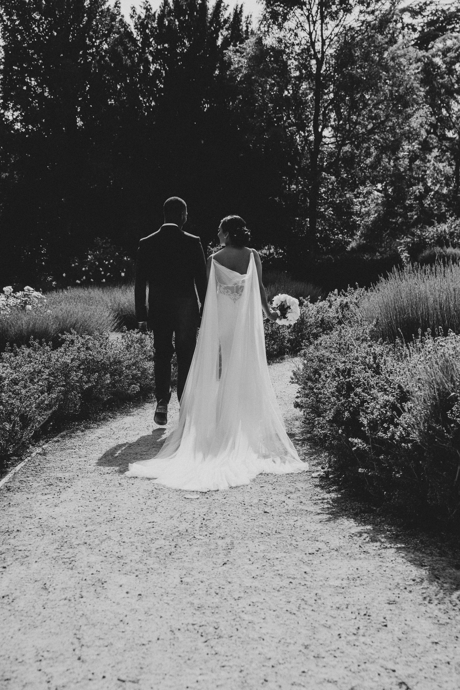 Black and white photo of bride and groom walking hand in hand down Langtons House garden path lined with lush greenery.