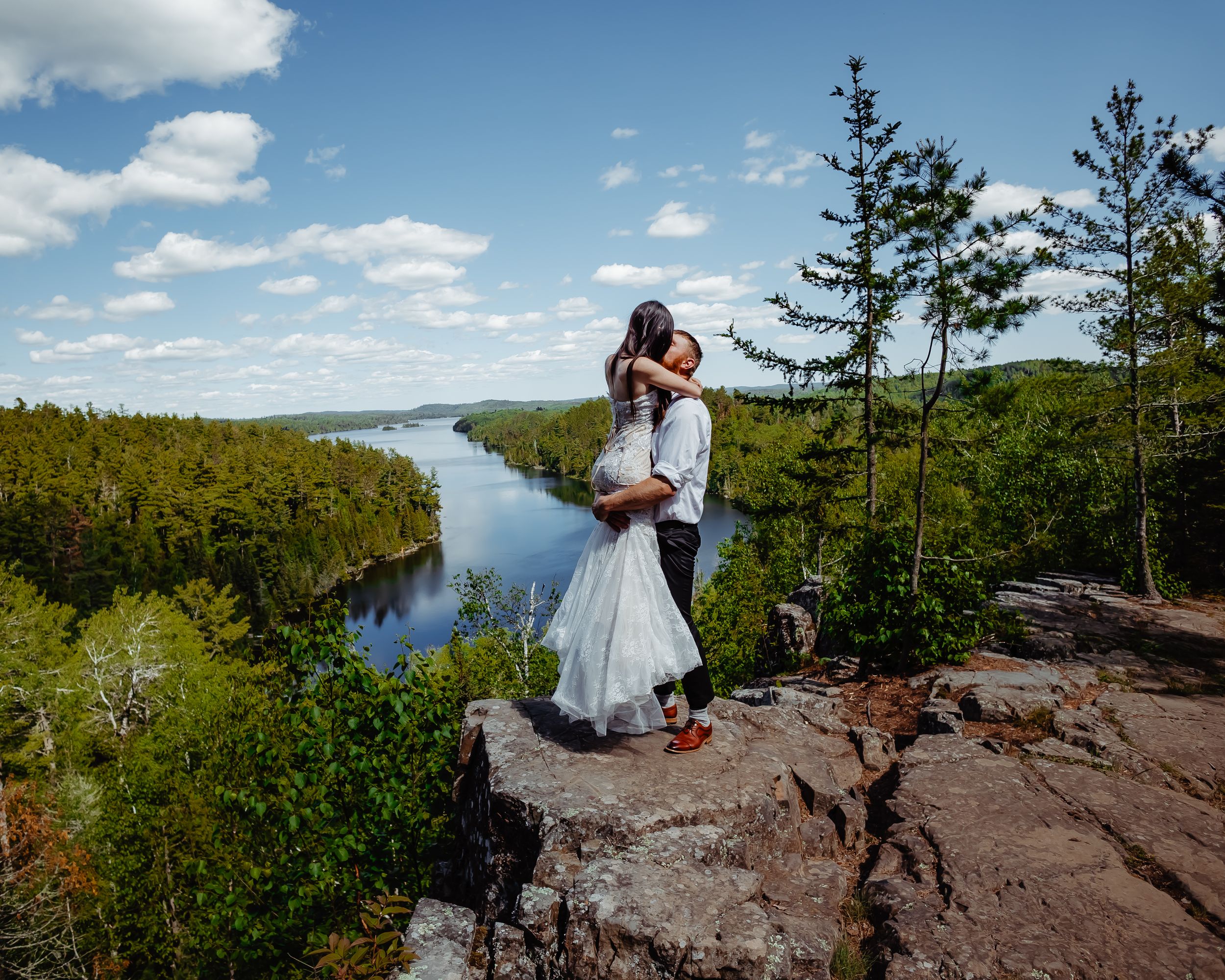A couple in wedding attire shares a romantic moment on a scenic cliff overlooking a lake surrounded by lush forests.