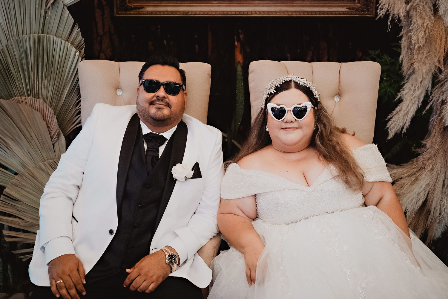 A bride and groom wearing sunglasses pose together at their wedding while sitting in cream-colored chairs.