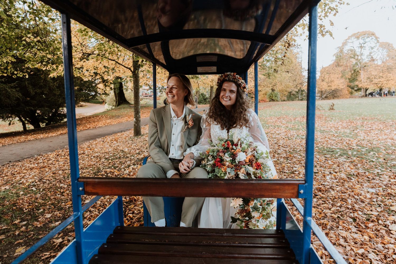 Newlywed couple sits on blue trolley surrounded by fallen autumn leaves during outdoor wedding photo session.
