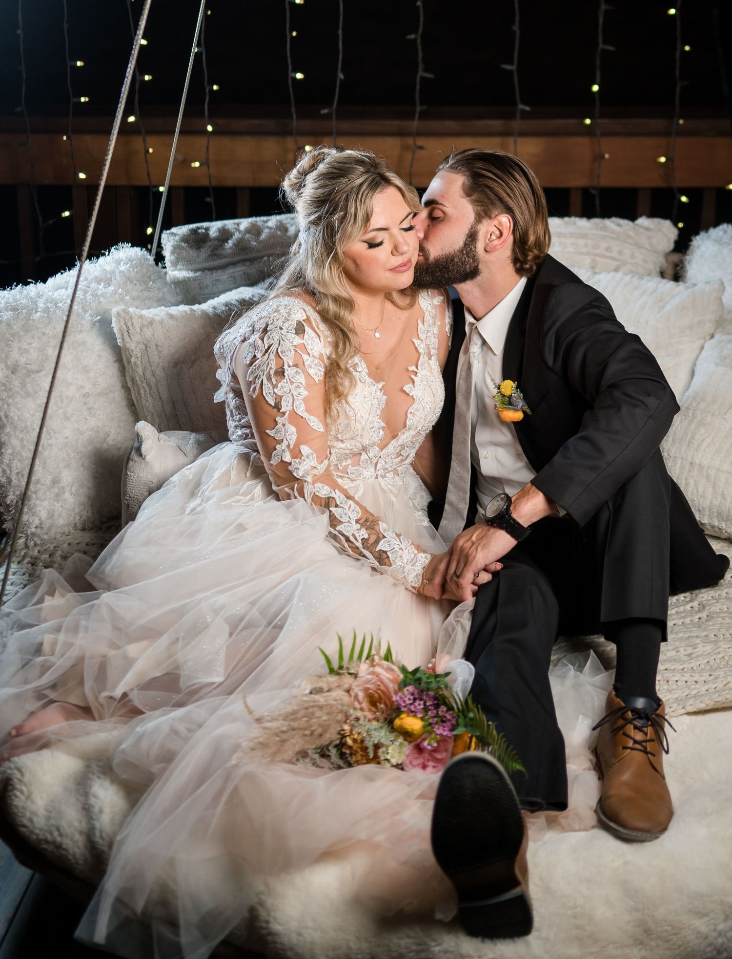 Couple shares intimate moment on plush white seating with bride wearing lace sleeve gown and holding colorful bouquet.