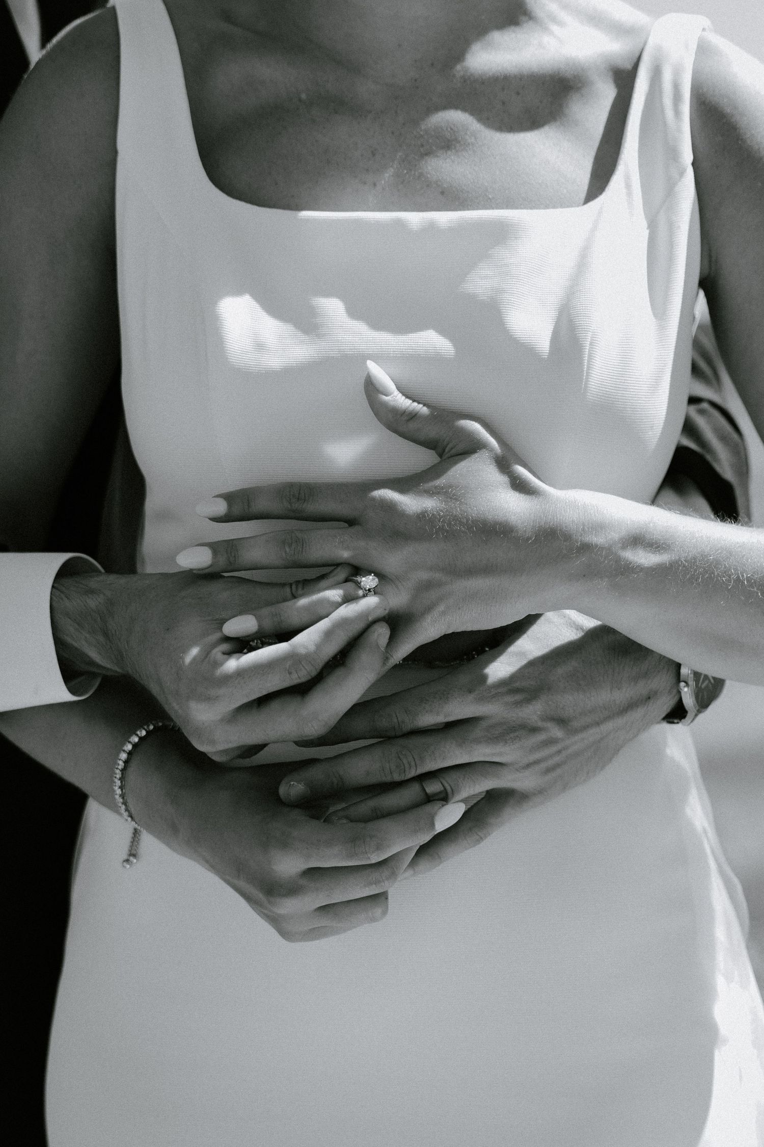 Black and white close-up of hands crossed over a white dress, creating an intimate self-embracing pose.
