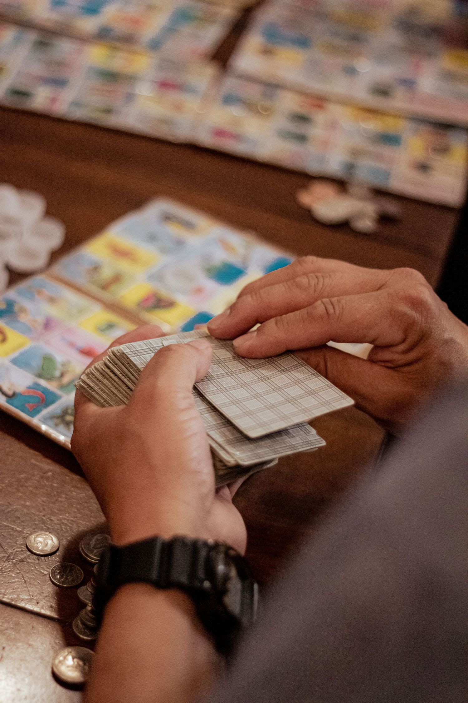 Close-up view of hands holding playing cards with colorful board game elements spread on wooden table surface.