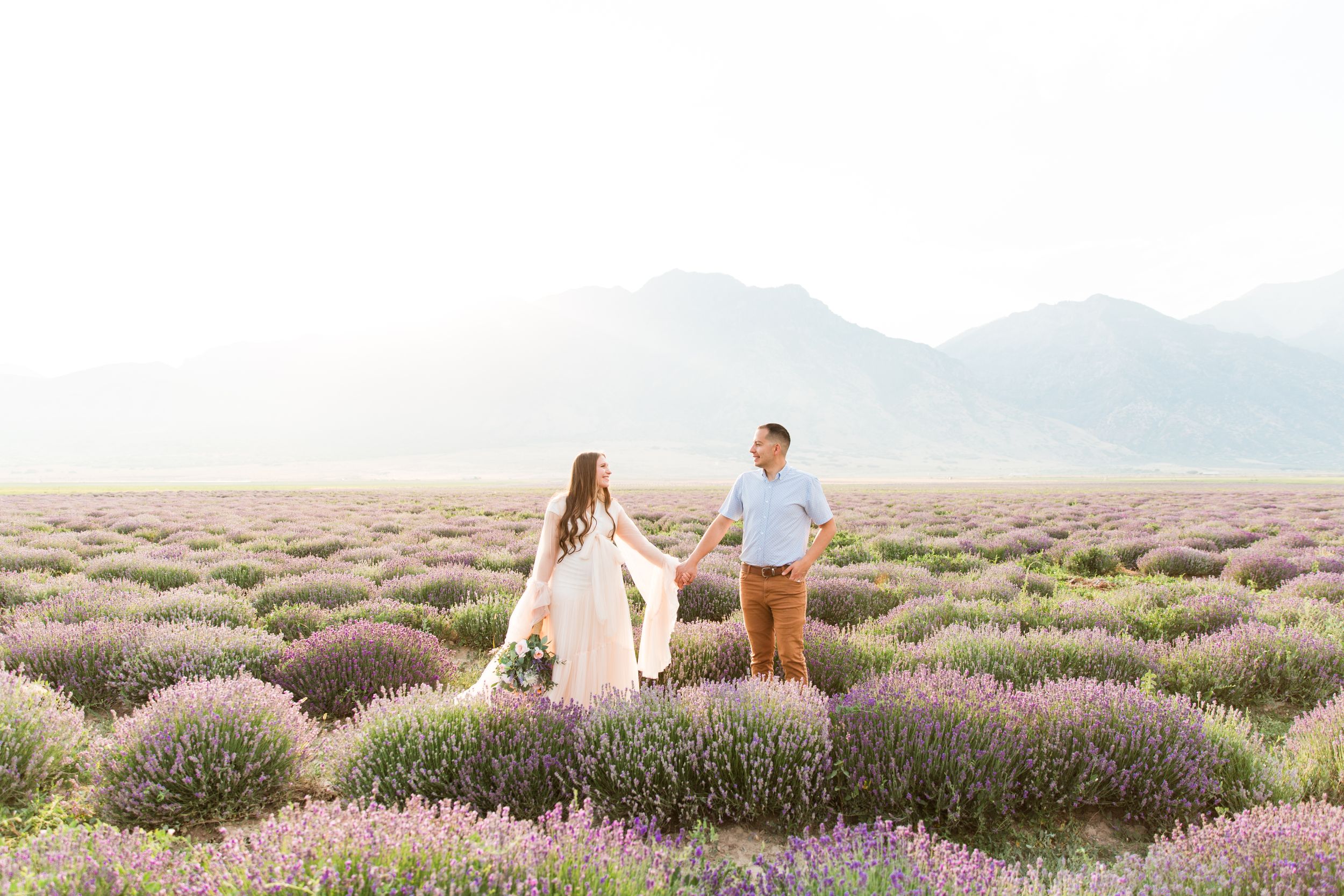 Serene maternity shoot in lavender field, backlit by sunset with mountain views. Mom-to-be in flowy dress holding flower