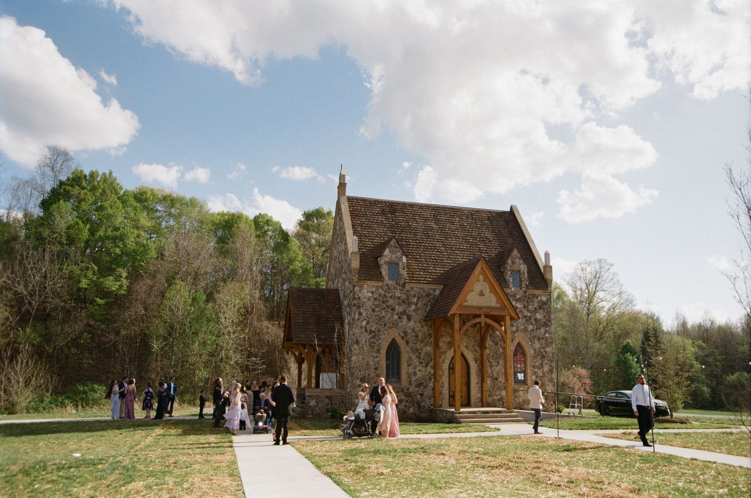 Historic stone chapel with wooden entryway stands among green grounds as visitors explore the scenic outdoor space on a sunny day.