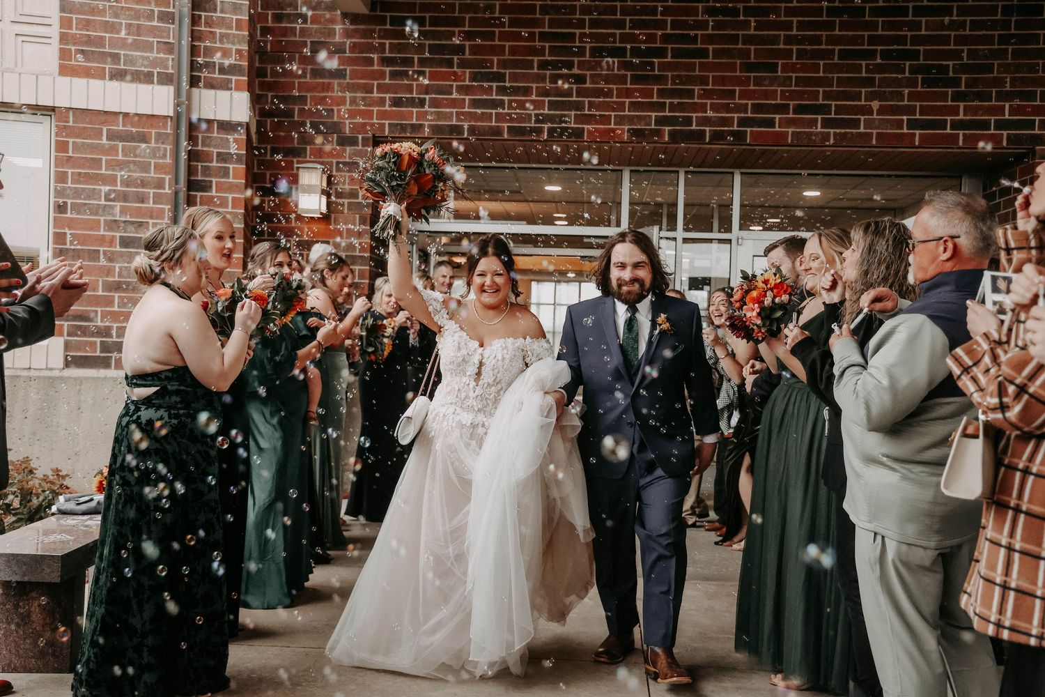 Wedding party exits venue as guests throw confetti during joyous celebration with brick wall backdrop.