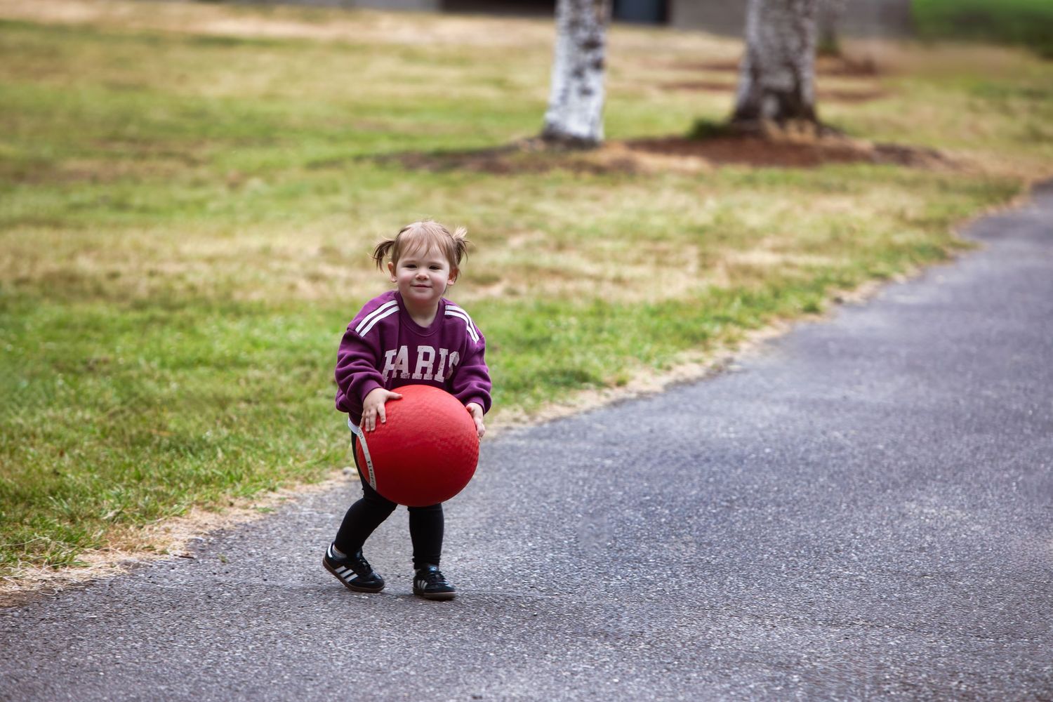 Small child in a winter jacket holds a large red bouncy ball while standing on a paved pathway outside.