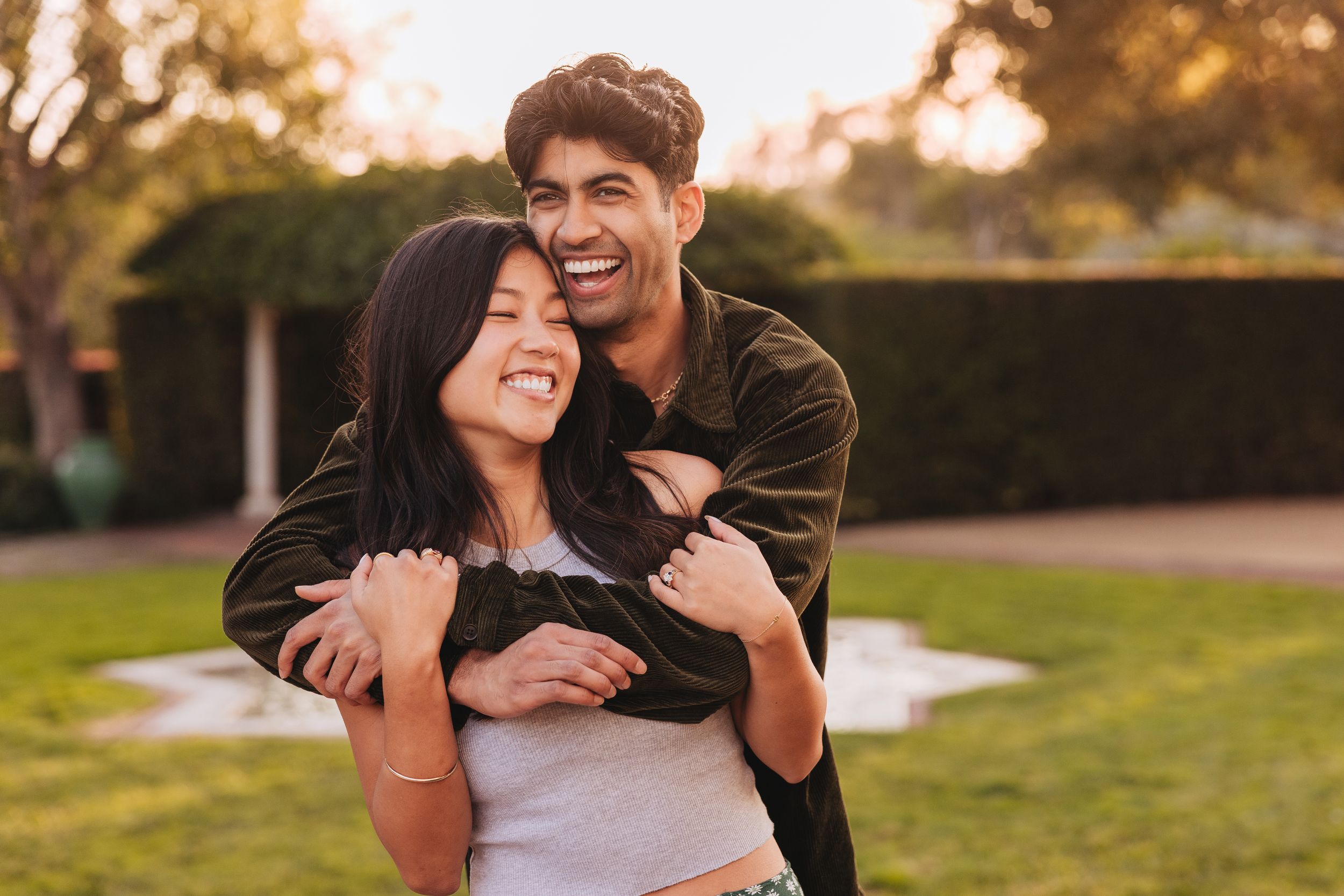 A happy couple embraces and laughs together outdoors during golden hour in a park setting.