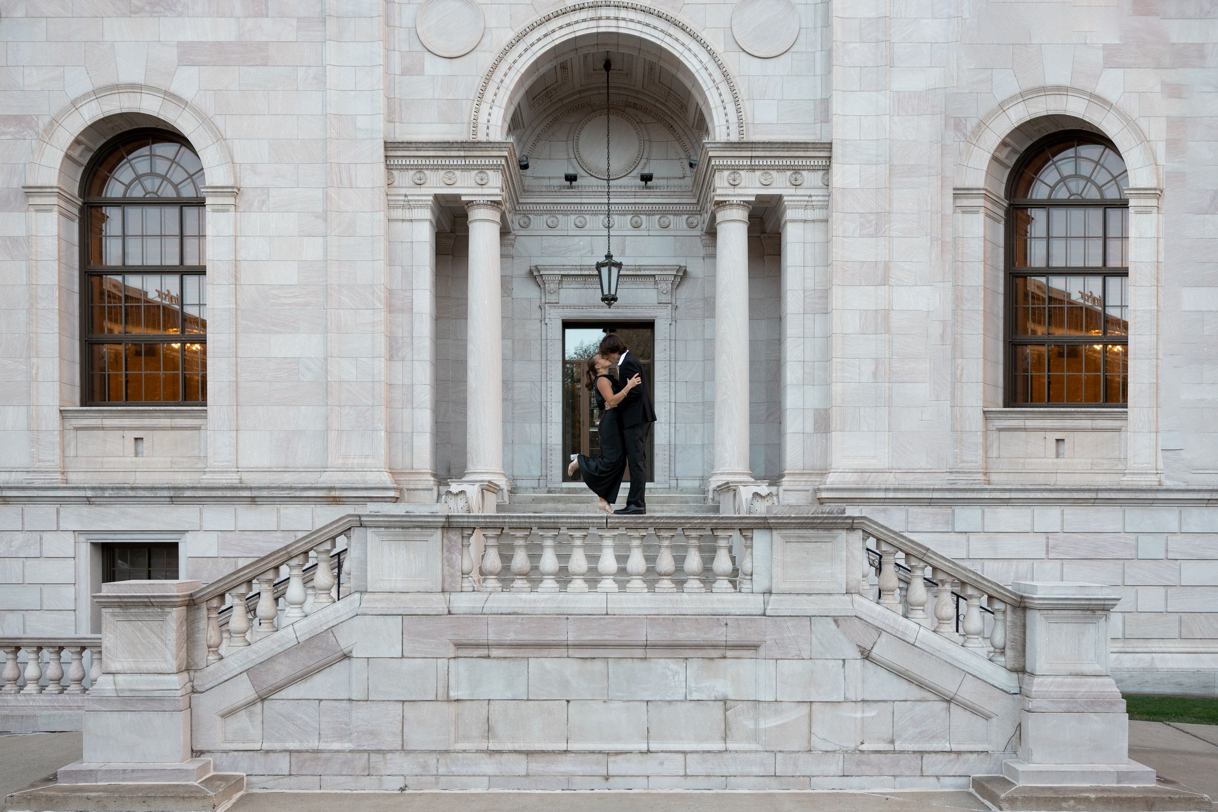 Person in graduation cap and gown standing on steps of grand stone building.