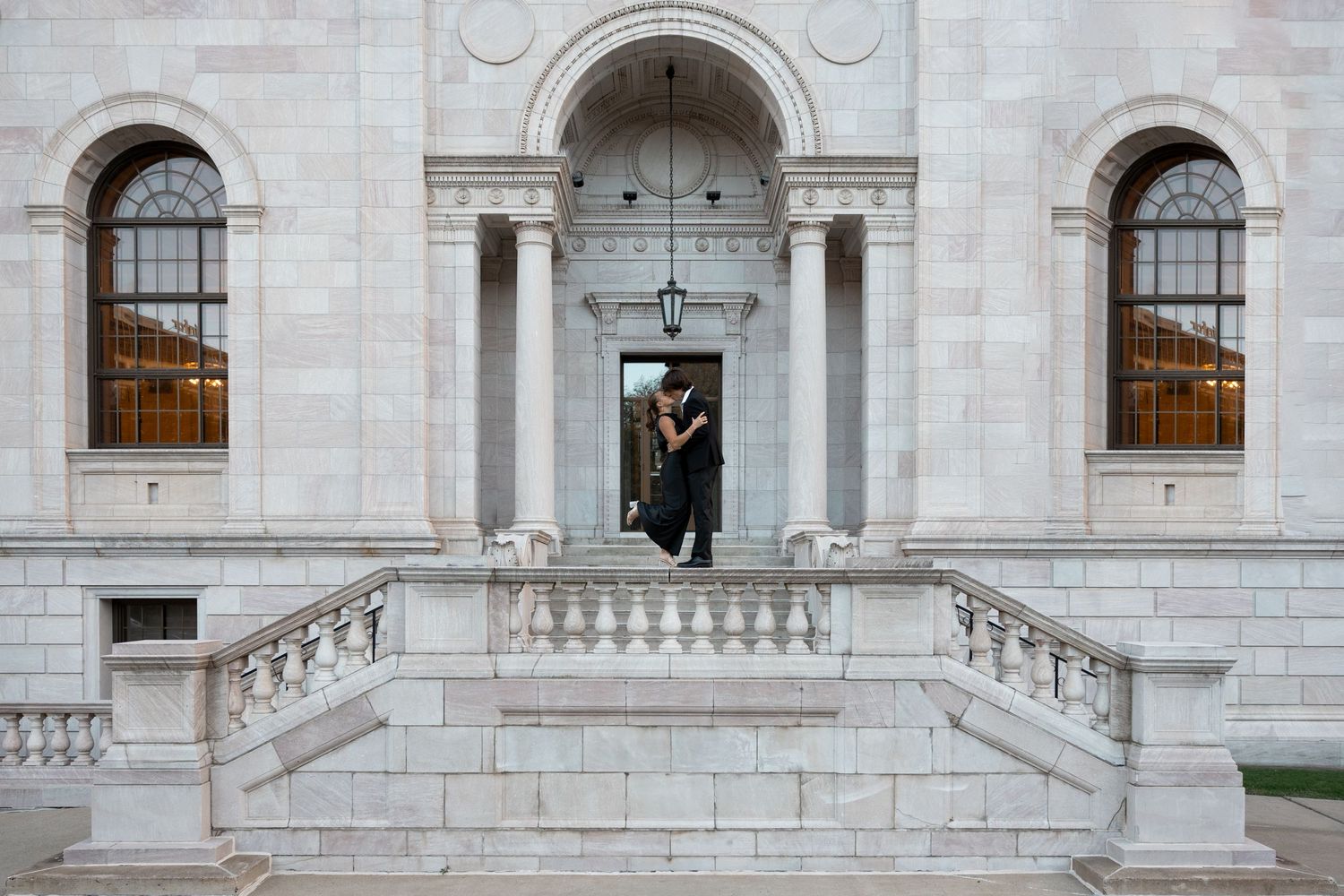 Person in graduation cap and gown standing on steps of grand stone building.