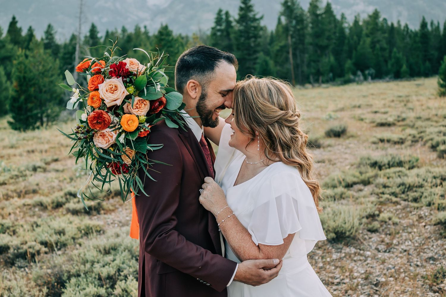 Bride and groom embrace in a field in the Tetons surrounded by trees with bride holding a bouquet