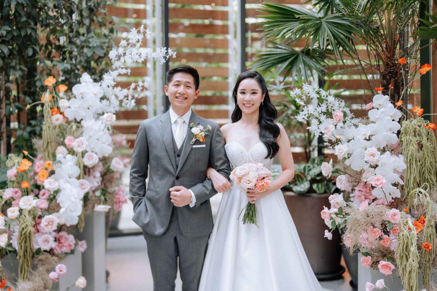 Wedding couple posing under floral arch with pink and white blooms and tropical greenery.