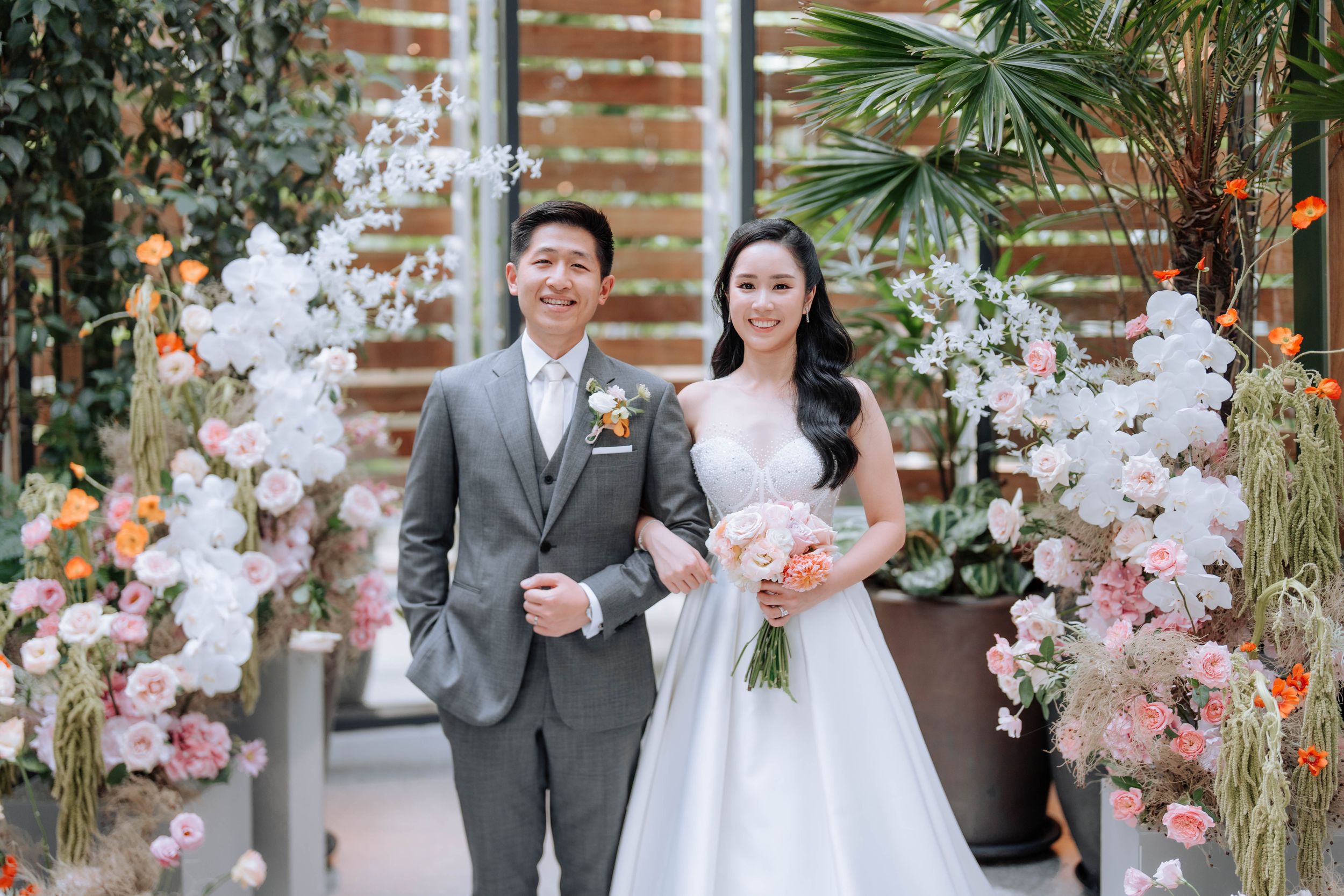 Wedding couple posing under floral arch with pink and white blooms and tropical greenery.