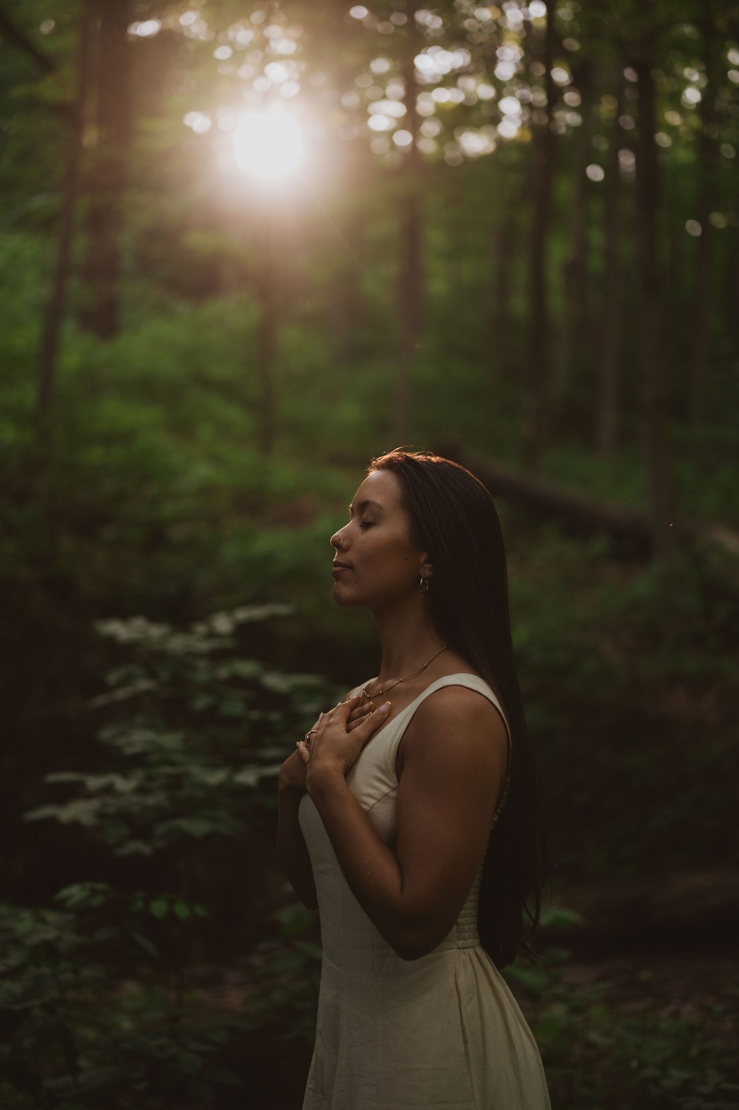 A woman in a white dress stands peacefully in a sunlit forest with eyes closed during golden hour.