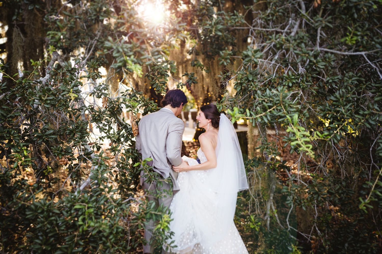 Romantic garden wedding scene with couple in white sharing an intimate moment backlit by golden sunlight through trees.