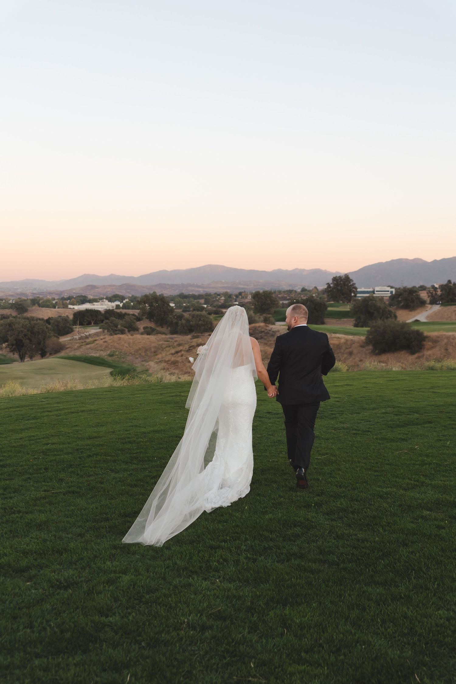 A wedding couple walks on a grassy hill overlooking mountains during sunset with the bride's veil flowing behind them.