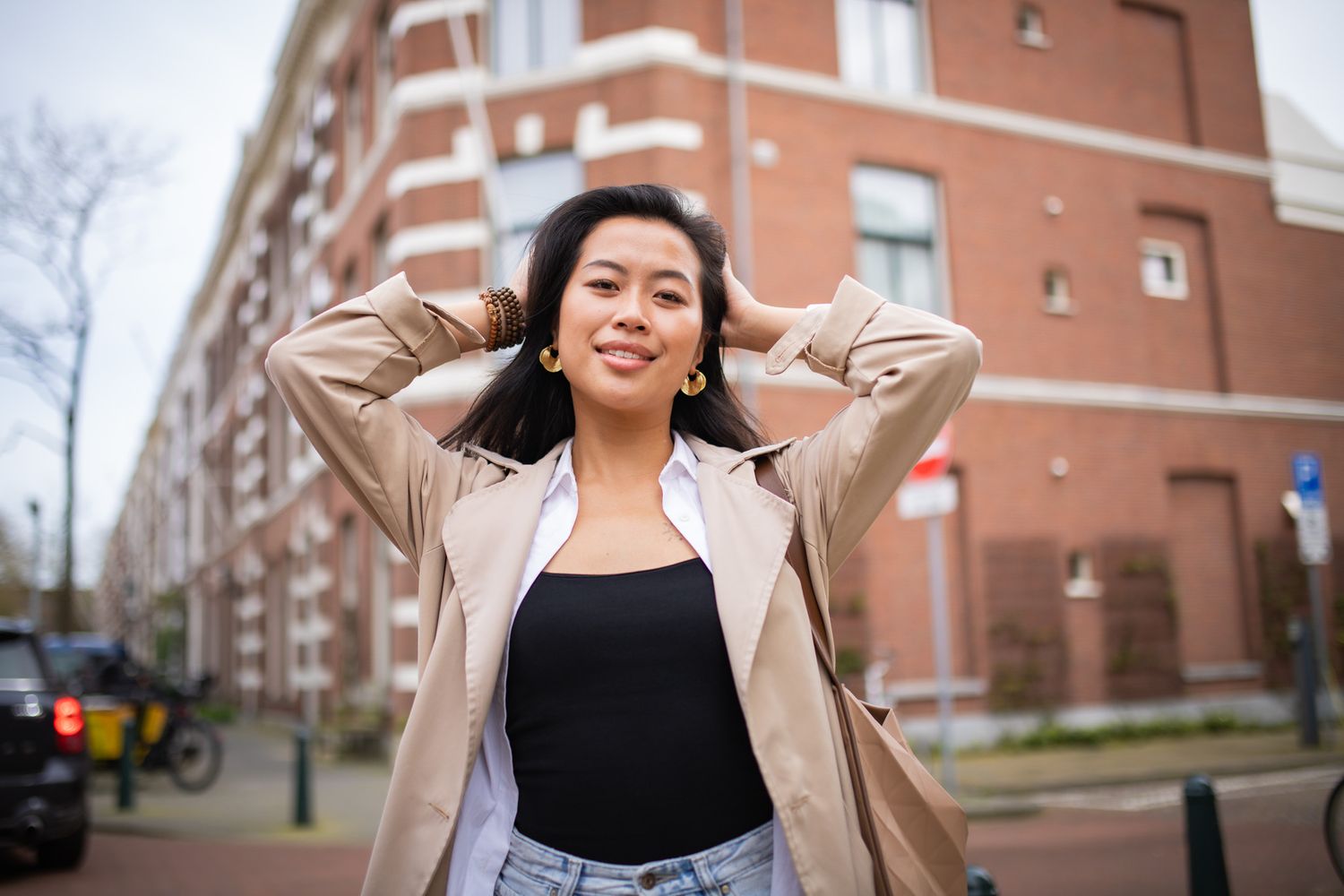 A person in a beige jacket and black top poses with hands behind their head on a city street with brick buildings.
