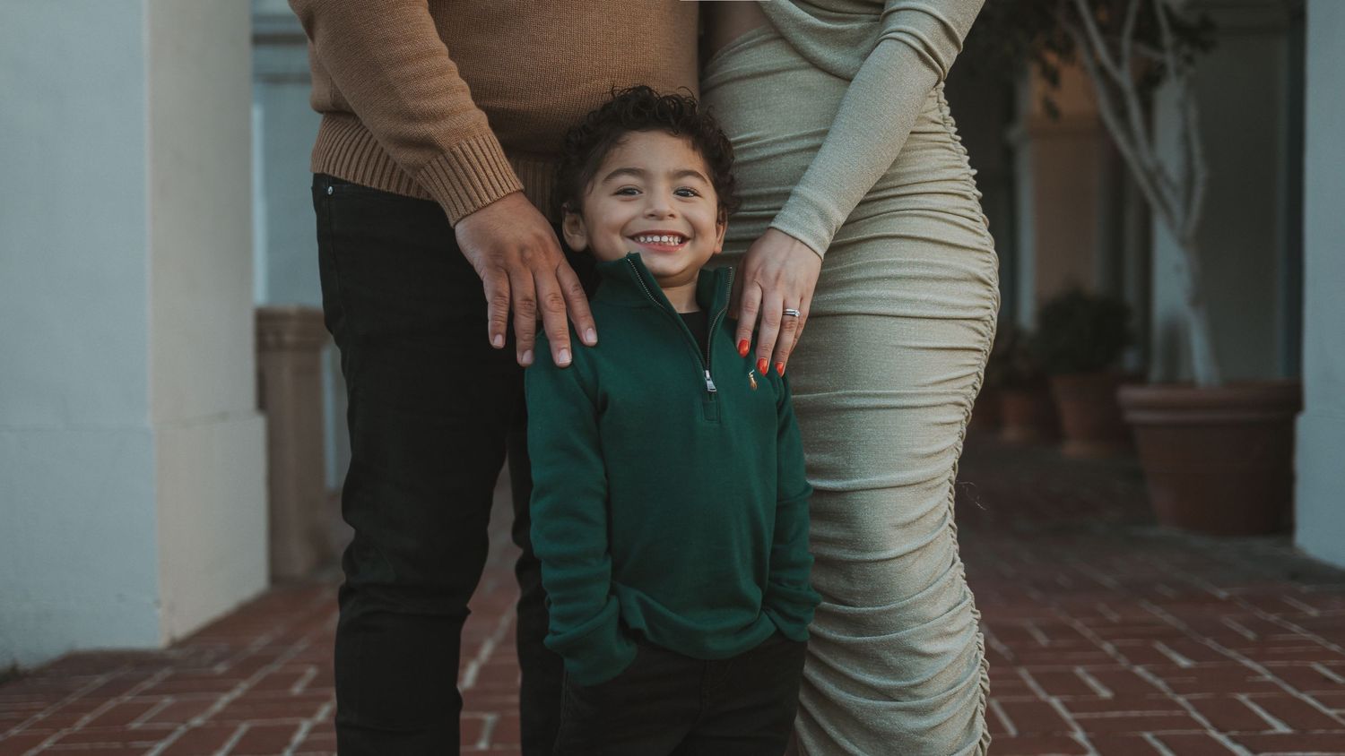 A young child in a green sweater stands between two adults on a brick patio with columns in the background.