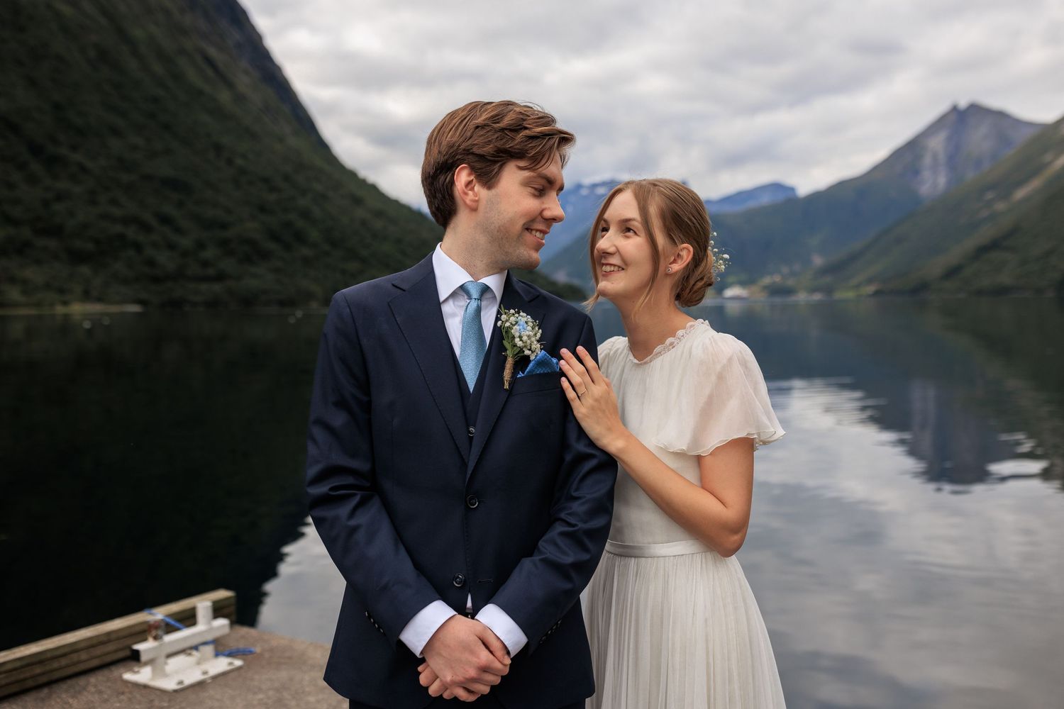 A couple shares a joyful moment by a scenic mountain lake in wedding attire.
